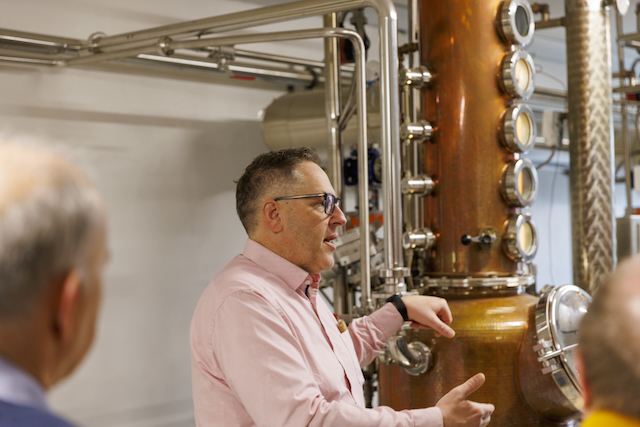 Ein Mann mit Brille, wohl ein Brennmeister, erklärt vor einer grossen, glänzenden Kupfer-Destillieranlage etwas. Im Vordergrund sind die Köpfe von zwei Zuhörern zu sehen. Die Anlage deutet auf den Macardo Brenn-Kurs hin.